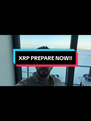 A selfie-style video featuring a man in a light-colored shirt standing in front of a large window with a cityscape and water in the background. A rectangular text overlay in teal and pink reads 'XRP PREPARE NOW!!'.