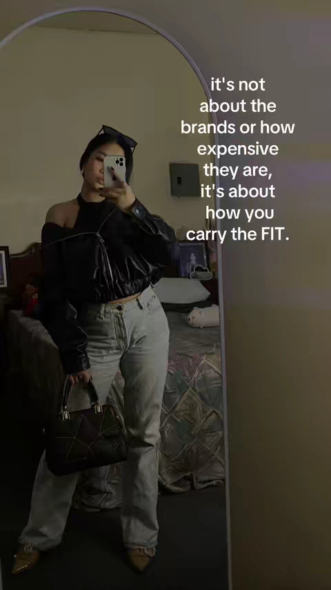 A person is taking a mirror selfie. The person, with a neutral expression, is wearing a black top, sunglasses on their head, and light colored jeans. Text overlays the image, and the mirror has a clear reflection of the setting around them.