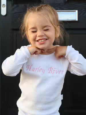 A young child, likely a toddler, stands smiling in front of a black door. The child appears to be female with blonde hair, and she is wearing a white long-sleeve shirt with the name "Harley River" printed in pink across the chest. She is holding the collar of her shirt and has her eyes closed.