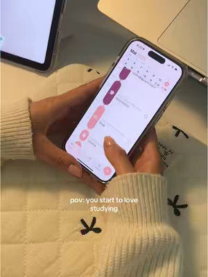 A person is holding a smartphone displaying a calendar app with a detailed study plan on a white patterned surface. The person's hands and the smartphone are the primary focus, with a laptop and tablet visible in the background in a natural light setting.