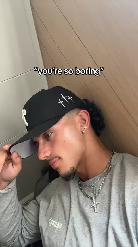 A young man with dark curly hair, wearing a backwards black cap with a white 'C' and white crosses on it, a gray long-sleeved shirt, and a silver necklace with a cross pendant. He is in front of what appears to be a plain off-white wall and wooden brown side wall.