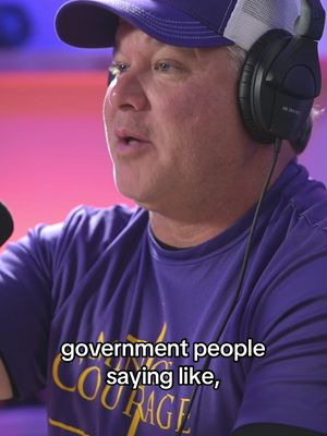 A man with light skin wearing a purple baseball cap with a white mesh back and a purple t-shirt. He is wearing black over-ear headphones. The background is blurred with shades of pink and blue. Text overlay on the image reads, 'government people saying like,'