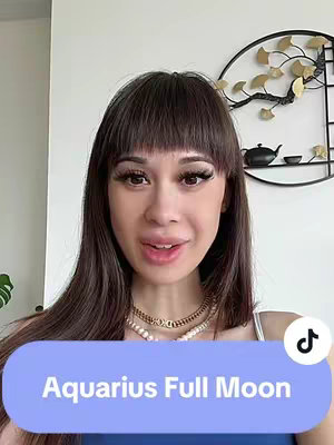 A woman with long brown hair and light skin is centered in the frame. She is wearing a light colored top and a necklace. Behind her is a decorative piece with a circular design. At the bottom of the frame, there's a purple banner with white text that reads 'Aquarius Full Moon'. A TikTok logo sits to the right.