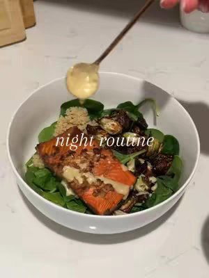 A close-up shot of a white bowl filled with a colorful salad. The salad contains a piece of grilled salmon, green leafy vegetables, cooked grains, and roasted vegetables. A golden spoon hovers above, drizzling a creamy sauce over the dish. The overall composition is well-lit, with a clean aesthetic.