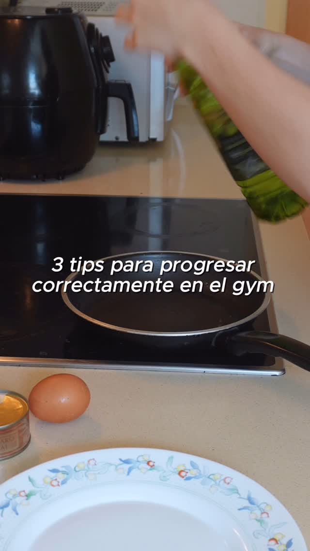 A person is pouring oil from a bottle into a frying pan on a stovetop. Nearby are an egg, a can of tuna, and a plate with a floral design. Text overlays read "3 tips para progresar correctamente en el gym".