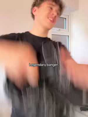 A young man with light skin and curly brown hair is captured from the chest up. He is smiling with a slightly open mouth, and his eyes are looking towards the viewer. He is wearing a black t-shirt. The background appears to be a neutral-colored wall with a white-framed window visible on the right. In the foreground, the image is slightly blurred to highlight the subject against the background. Text that reads Legendary banger is superimposed on the image as well.