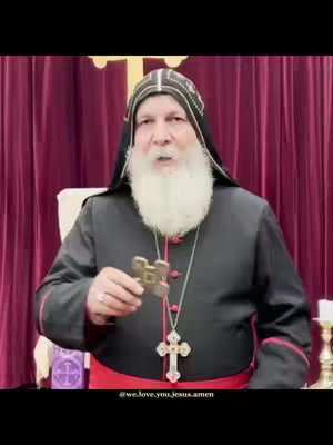 An adult male with a white beard and dark clothing giving a prayer. He's holding a crucifix and wearing a religious head covering with a cross in the background.