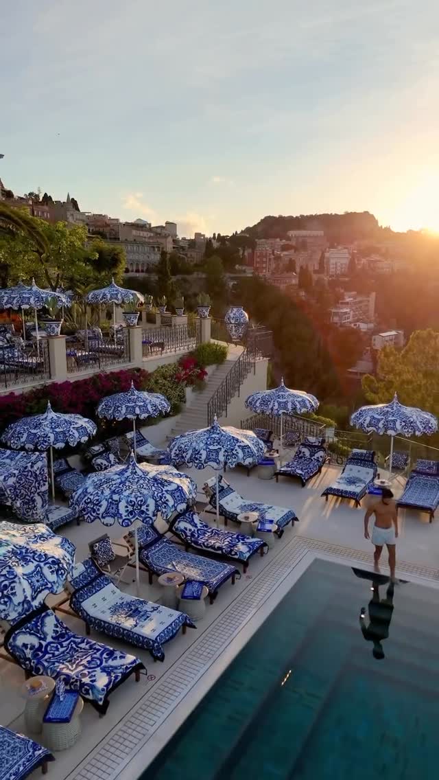 An elevated view of a luxury hotel pool area. The scene is set at golden hour with sunlight setting over a town in the distance.  The foreground features a pool with a man standing at the edge.  The pool is surrounded by lounge chairs and tables with blue and white patterned umbrellas.  The design is repeated on the lounge cushions and the table tops. Behind the pool area is a terraced area with more seating, greenery, and a set of stairs.
