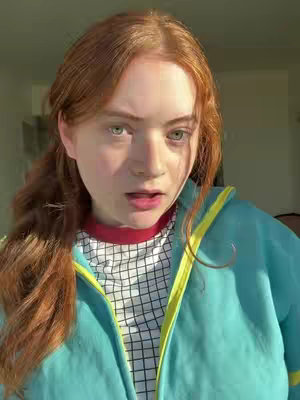 A young woman with medium-length auburn hair that is parted on the side looks at the camera with a slightly open mouth and questioning expression. She is wearing a white and black checkered shirt with a red collar under a turquoise jacket with a yellow trim. The shot is illuminated with a warm light, likely from a window.