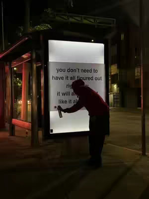 A night scene shows a person in a red hoodie and dark clothing, standing by a lit bus stop. The person is spraying a message onto a bright, white signage. The message appears to be a motivational quote. The background includes cityscape elements, with streetlights illuminating the scene.
