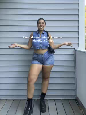 The image shows a woman standing outdoors, posing in front of a wall of gray siding. She looks at the camera with both arms outstretched, and is wearing a denim crop top and shorts with black boots. Text overlay is present at the top, and the overall lighting is bright.