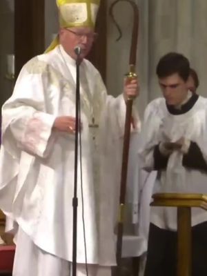 An older man in a white religious garment and yellow hat is speaking into a microphone. He is holding a staff and there is another person in the background with a white garment.