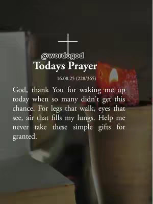 The image is a vertical, slightly blurred photo featuring a warm, muted color palette. In the foreground, there's text with the header 'Todays Prayer' positioned below a white cross symbol. The text is a prayer thanking God for a new day and the simple gifts of life. Background elements include a candle's soft glow and indistinct shapes, implying a devotional setting.