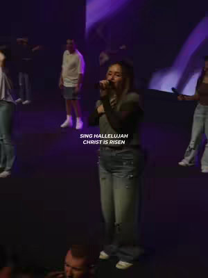 A vertical shot shows a woman singing on a stage with other people and purple lighting. The main subject is a woman centered, singing into a microphone. She is wearing a dark long sleeve shirt and faded jeans. Behind her, there are several people; some are also holding microphones. The background consists of a large screen with abstract shapes and patterns in shades of purple.