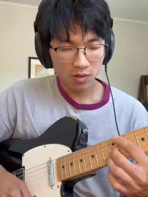 A young man with glasses and headphones is seen playing an electric guitar. The guitar is black and white, and the focus is on his hands and the fretboard. He's seated in a room with a painting in the background.