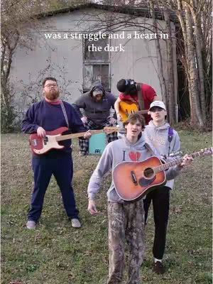 A group of five men standing outdoors in front of a building. Four of them are holding guitars, with one man at the front of the center with the guitar and another in the back, holding a drum.