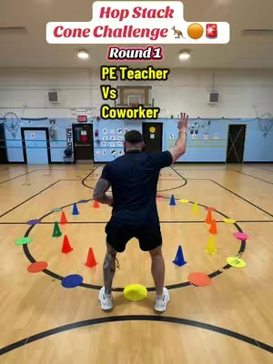 A man stands in an indoor basketball court setting, facing away from the camera. He is surrounded by colorful cones and circular mats, arranged in a circle on the court. The scene is well-lit, with a focus on the man and the challenge layout.