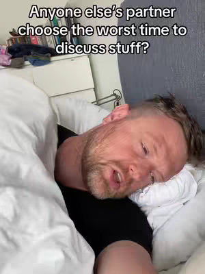 A man is lying in bed, appearing to be trying to sleep. Text overlayed on the image reads, 'Anyone else's partner choose the worst time to discuss stuff?' His head rests on a pillow, and he is covered with a white blanket. Behind him, a part of a cluttered nightstand is visible.