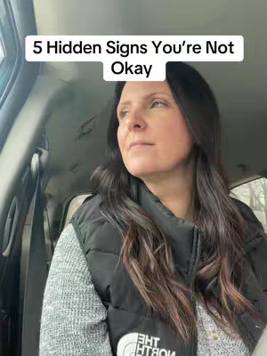 A woman is seated inside a vehicle, looking pensively towards the right with a neutral expression. Above her head, a white rectangular text box reads "5 Hidden Signs You're Not Okay". The background includes the car's interior and some glimpses of the outside environment, suggesting it might be daytime. The woman has long dark hair and is wearing a gray sweater under a black vest. The focus is on the woman's face and upper body.