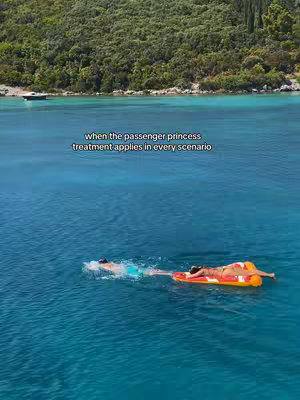 An aerial view of a turquoise ocean with two people swimming towards an orange and white inflatable raft. Green trees and bushes line the shore in the background.