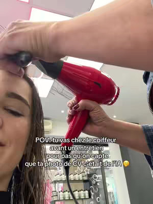 A woman is at a hair salon. Her face is in the lower left quadrant of the frame, with her eyes closed. A person is holding a red hair dryer directly in front of her, focusing on her hairline. Text overlays appear in the bottom center.