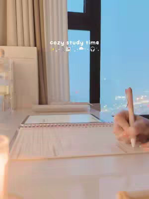 The cover image shows a cozy indoor scene focusing on a person studying. The scene includes a person writing with a pen on a notebook laid on a table, with a window with a city view behind the person. The lighting is soft, and the color palette is largely neutral with a touch of blue from the outside. The image is framed to highlight the study setup with a minimalist aesthetic.