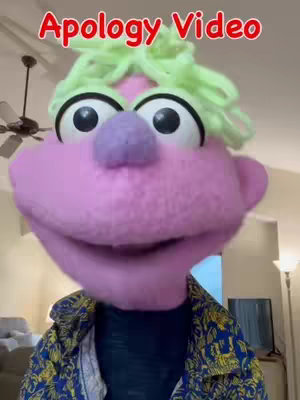A puppet with a large pink face, big black eyes, a purple nose, and light green, spiky hair is the central focus. The puppet is wearing a blue shirt and a colorful jacket. The background shows a room setting with a ceiling fan and a distant TV.