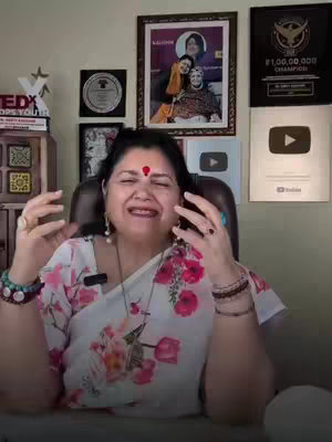 The cover image shows a woman seated indoors, likely in front of a desk or bookshelf. She appears to be in her 40s or 50s, with dark hair, a red bindi on her forehead, and a white and floral-patterned top. She is smiling and gesturing with both hands, suggesting she is animated while speaking. Behind her are framed pictures, awards, and a YouTube play button, indicating success or recognition. The lighting appears to be natural, casting a pleasant glow on the scene. The overall composition and style suggest a video focused on spiritual or astrological content.