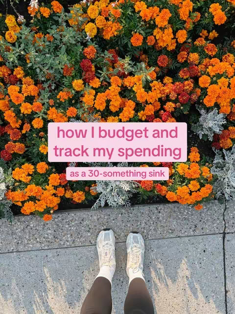 A photo taken from a low angle features a person's feet and legs standing on a light gray stone path. The person is wearing white sneakers and dark leggings, with white socks peeking out above the shoes. In the background, a vibrant display of orange and yellow flowers provides a colorful, eye-catching backdrop. A rectangular overlay, colored in a pastel pink, is positioned in the middle of the image, and on top of the flowers, containing white text related to budgeting and personal finance.