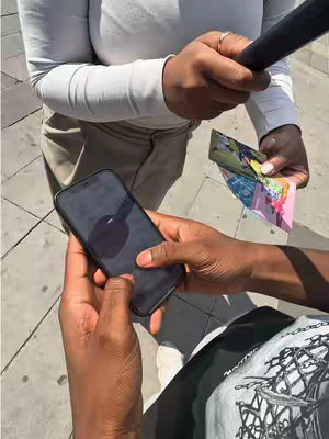 A candid overhead shot shows two people's hands holding a phone and some vibrant cards. The person in the foreground has darker skin and holds the phone, a black iPhone, displaying a dark screen. The other person holds a colorful set of cards. The background shows the pavement with sunlight on it..