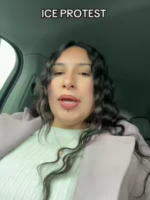 A woman is seated in the front seat of a car, facing forward. The image is a selfie-style shot. Text overlay 'ICE PROTEST' is present at the top. The woman has long black curly hair, wearing a white top and a light-colored jacket. The background shows the interior of the car.