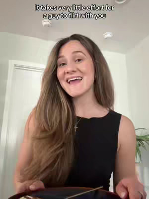 A woman with long brown hair smiles at the camera, holding a dark plate with food. Text overlayed above reads, 'it takes very little effort for a guy to flirt with you.' The background includes a white door and ceiling lights.