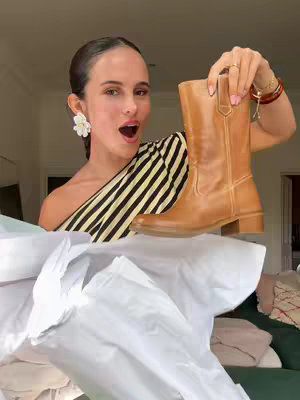 A woman with dark hair and fair skin stands indoors, holding up a light brown cowboy boot. She's wearing a black and yellow striped top and a large white flower earring. Her mouth is open in an expression of excitement. The background shows a glimpse of a room with white walls and hints of furniture and soft items. The lighting is natural and soft.
