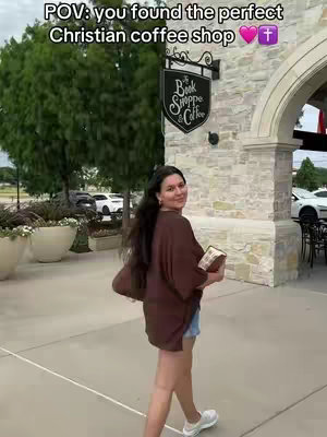 A woman with long dark hair is walking toward the camera, holding a book and smiling. She's in front of a brick building with a sign that says 'Book Shoppe & Coffee.' The sky is bright, and there are trees and parked cars in the background.
