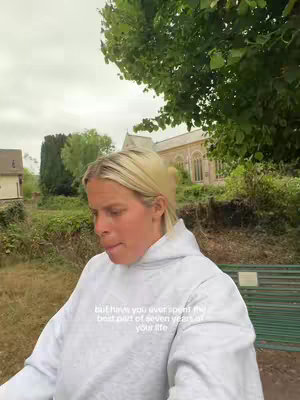 A woman with blonde hair wearing a light gray hoodie stands outdoors. Soft focus blurs the background, featuring greenery, a building, and the overcast sky. Text overlayed suggests a reflective tone.