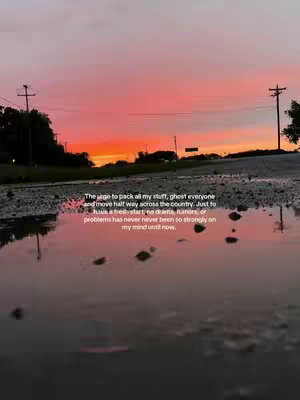 A photograph of a road with a puddle reflecting a colorful sunset. The sky is painted with shades of orange and pink. The puddle has a slightly reflective surface, and on the horizon, there are power line poles. The image is composed to capture the sunset and the reflection in the water.