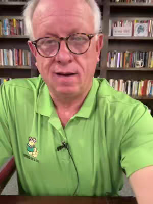 A middle-aged white man with glasses and short, graying hair is centered in the frame. He wears a light green collared shirt with a small cartoon frog emblem on the left chest. The background is a blurred bookshelf filled with books.