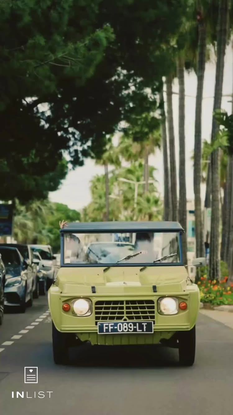 A light green vintage car drives down a tree-lined street. Other cars are visible behind it, and palm trees line the sidewalk. The image is bright and has a slight vintage feel.