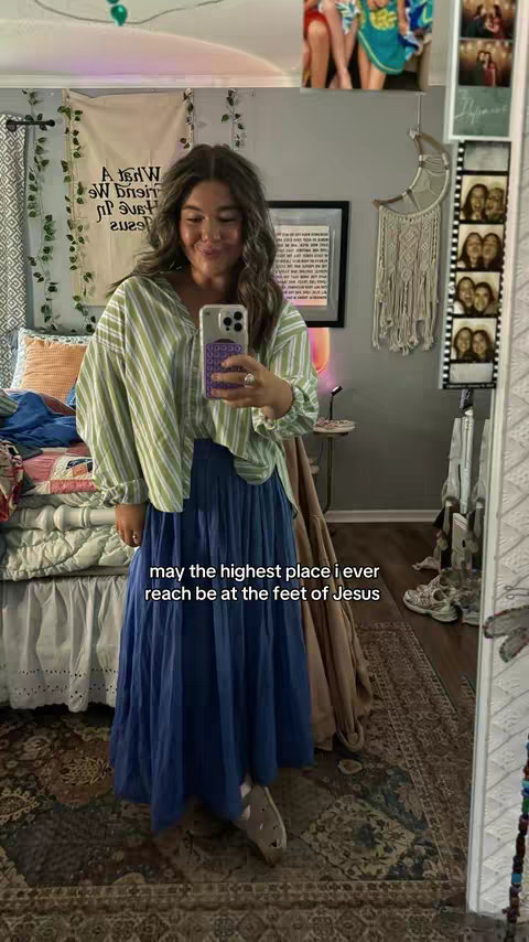 A woman is shown in a mirror selfie, dressed in a casual yet stylish outfit. She is smiling and appears to be in her bedroom, with various decorative elements and personal items visible in the background. The primary lighting seems to be natural, providing soft illumination. The color palette is a mix of soft, muted tones with occasional bursts of brighter hues from the clothing and room decor.