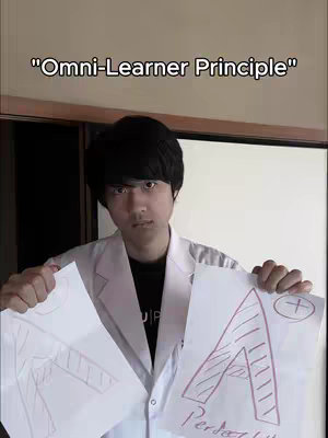 A young man in a white lab coat holds two paper illustrations. The background shows a plain wooden surface.