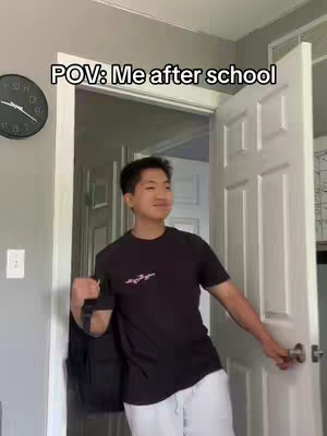 A young man, appearing to be of East Asian descent, stands in a doorway with the white door open, leaving an inside room entrance visible in the background. He is smiling slightly and holding a black backpack with one hand, he is also holding the white door open with the other hand, the room behind him is partially visible and softly lit. The background contains a wall clock. Text overlayed on the image reads 'POV: Me after school'.