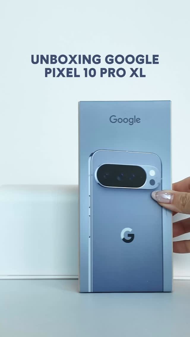 The image shows a person holding a Google Pixel 10 Pro XL box, partially obscuring a light-colored background on which a white, rectangular object is partially visible. The box is vertically oriented. The top right corner of the box displays "Google" in a sans-serif font. The phone appears to be light blue. The lower left of the phone's box displays the "G" Google logo.