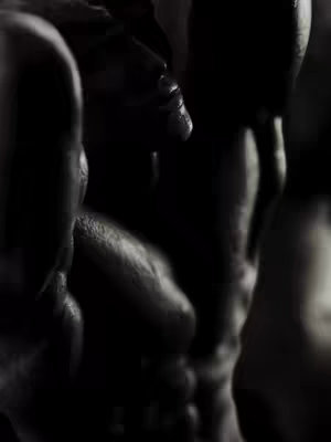A close-up, black and white image with multiple figures in the frame. The composition is dark, with the figures appearing to be partially obscured, adding a sense of mystery and depth. Highlights on the skin provide subtle contrast against the black background, emphasizing the body's form. The overall texture lends it a natural and potentially artistic feel.