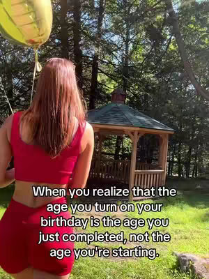 The image shows a woman from the back wearing a red athletic outfit, holding a yellow balloon, set against a backdrop of trees and a gazebo. Text overlayed on the bottom of the image reads: 'When you realize that the age you turn on your birthday is the age you just completed, not the age you're starting.'