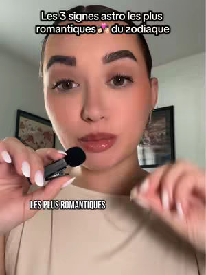 A woman with light skin, light brown hair, and makeup is centered in the frame, holding a small black microphone close to her mouth. Above her head, is text that reads 'Les 3 signes astro les plus romantiques' (The 3 most romantic astrological signs) with a pink heart emoji and a background image of paintings on a wall are slightly blurred. The overall setting appears to be indoors, with the woman positioned close to the camera, creating a close-up shot.