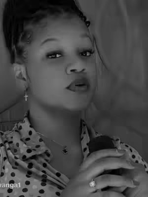 A black and white portrait of a woman with her hair up, wearing a shirt and holding a dark object, possibly a microphone. She has a cross earring and a ring. The background is blurred.