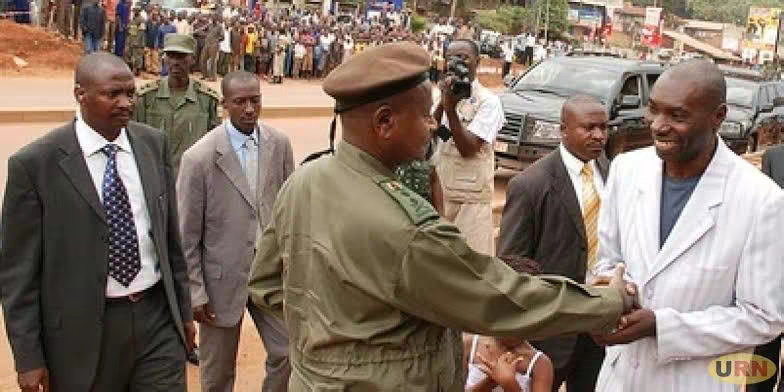 A photo shows a group of men outdoors. In the foreground, a man in a military-style uniform shakes hands with a man in a white suit. Other men in suits are nearby, and a crowd of people is visible in the background. The setting appears to be an outdoor event.
