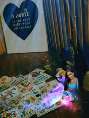 The image features a table with tarot cards scattered across. A woman is in the lower right corner. A framed artwork of a heart shape with the text inscription is visible. There is a draped curtain in the background.