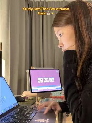 A woman is sitting at a desk with a laptop and a tablet in front of her. The tablet displays a timer app, and text overlays above her head which says "Study Until The Countdown Ends". The composition is framed from a side view, showing the woman's profile as she looks at the tablet display, the lighting is warm, the background is a blurred mix of desk items and a neutral-toned curtain.
