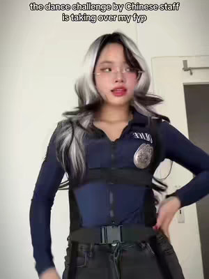 A young woman in a police outfit with black and white hair and glasses is posing for a video. The background appears to be a plain white wall with a door on the right.